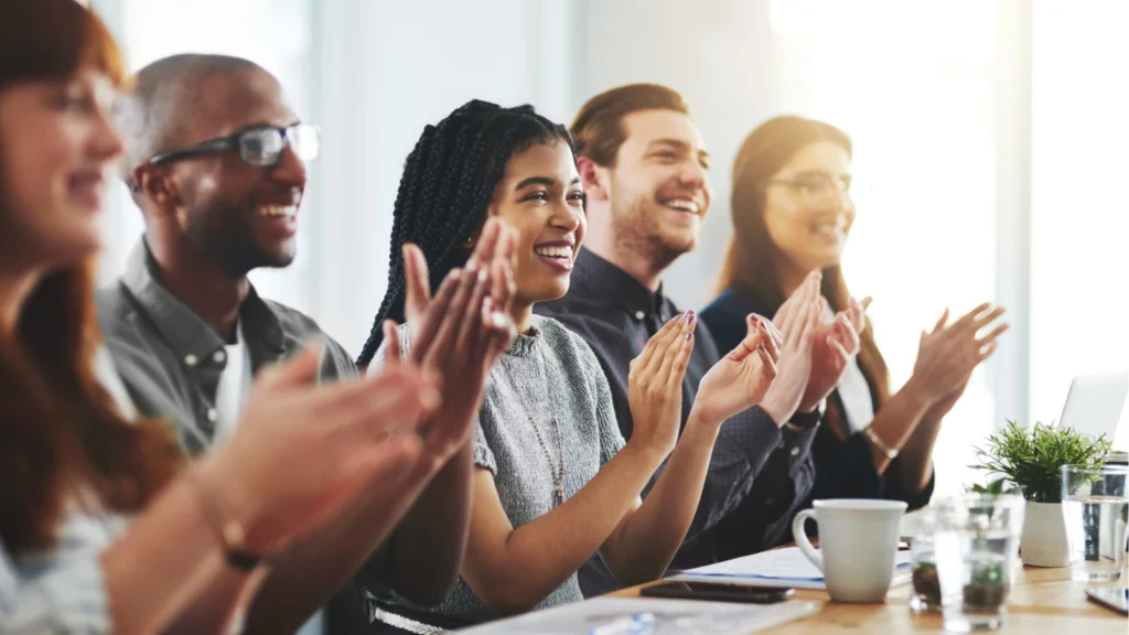 A diverse group of smiling colleagues sitting together at a conference table during a meeting, smiling and warmly applauding in appreciation.