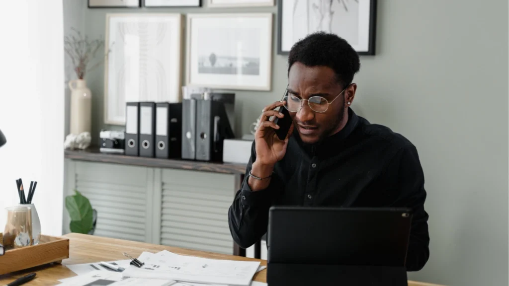 A focused man in glasses speaks on his phone at a desk, with a laptop, papers, and framed prints in a tidy office, appearing thoughtful and busy.