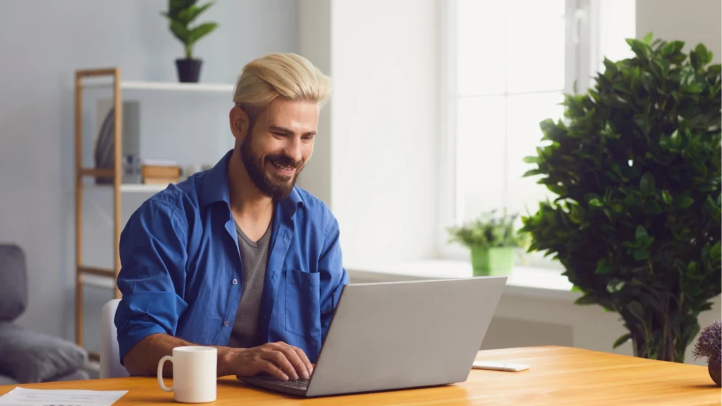 A smiling bearded man in a blue shirt works on a laptop at a sunlit desk, with a coffee mug and green plants creating a calm home-office setting.