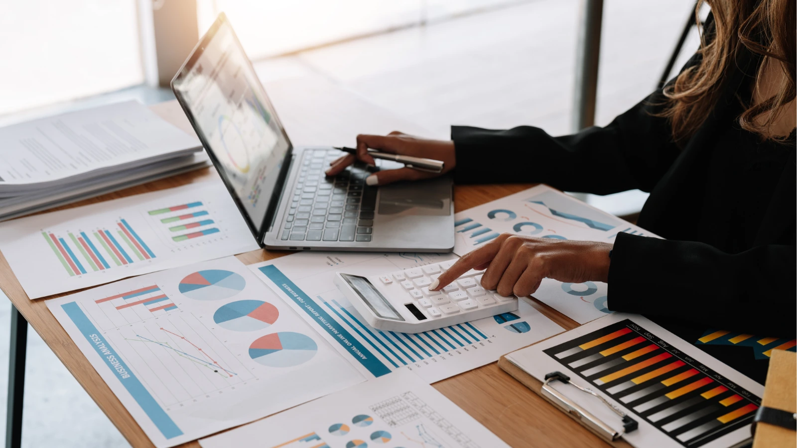 A business professional uses a laptop and points to charts, pie graphs, and a color-coded dashboard on a desk during focused data analysis.