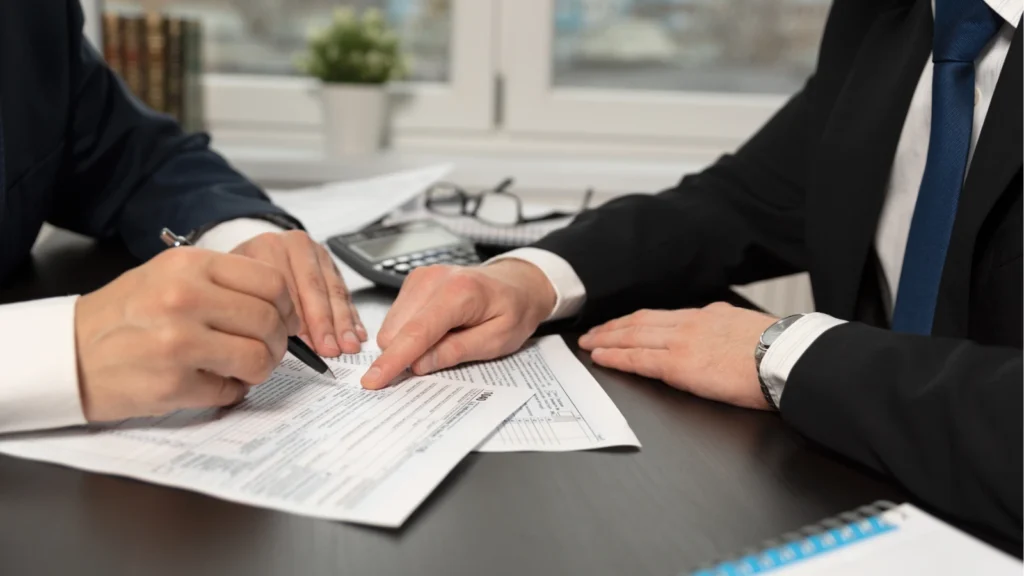 Two businesspeople review and sign paperwork at a desk, with a calculator nearby; the office scene conveys a focused, professional agreement.