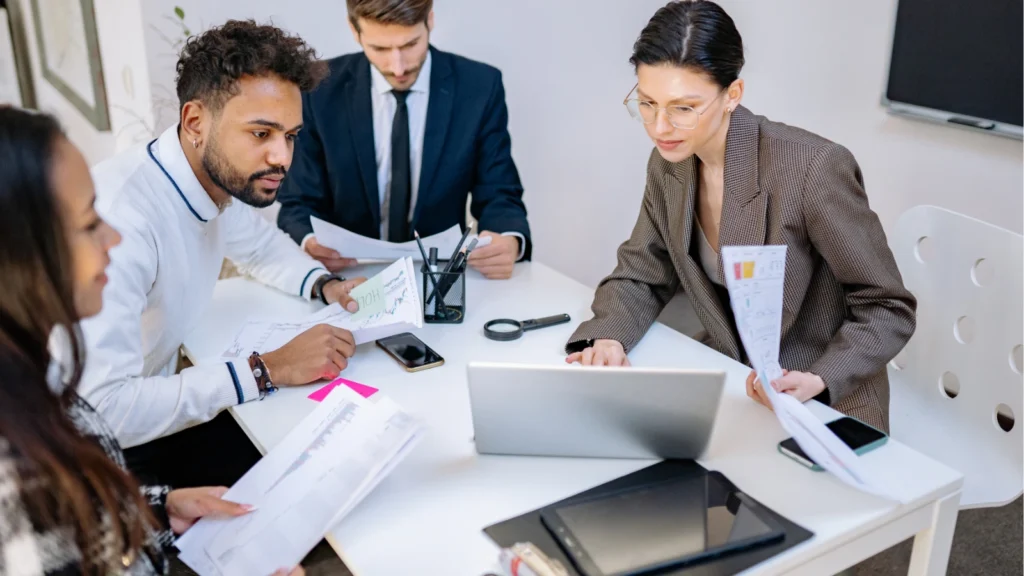 A team of corporate colleagues gathers around a conference table reviewing charts, documents, and a laptop.