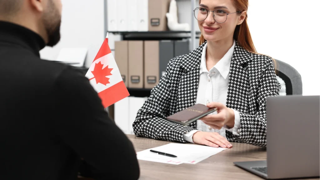 A woman in glasses hands a passport across an office desk with a miniature Canadian flag displayed on it.