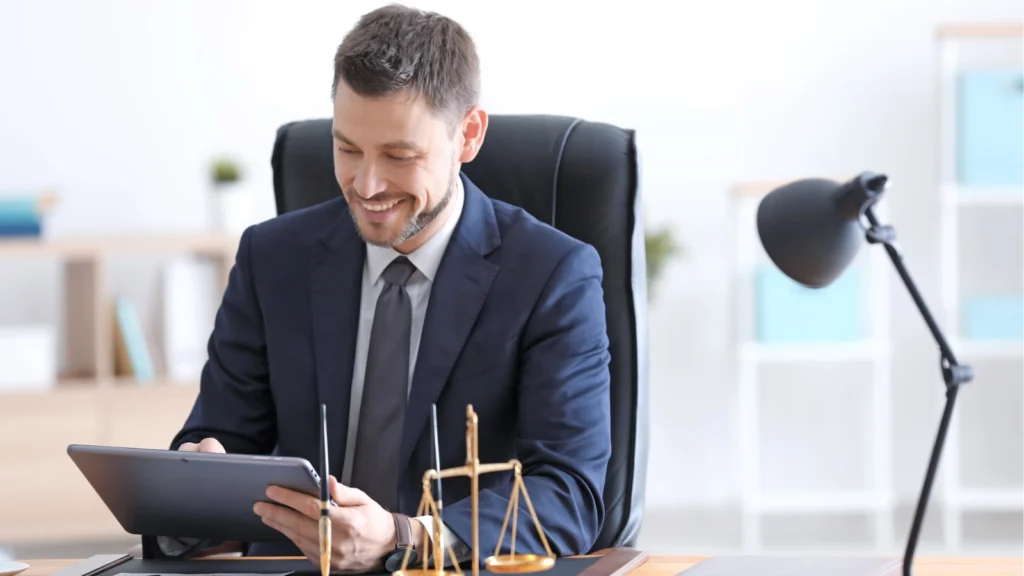 A smiling lawyer in a suit uses a digital tablet at his office desk next to a decorative scale of justice.