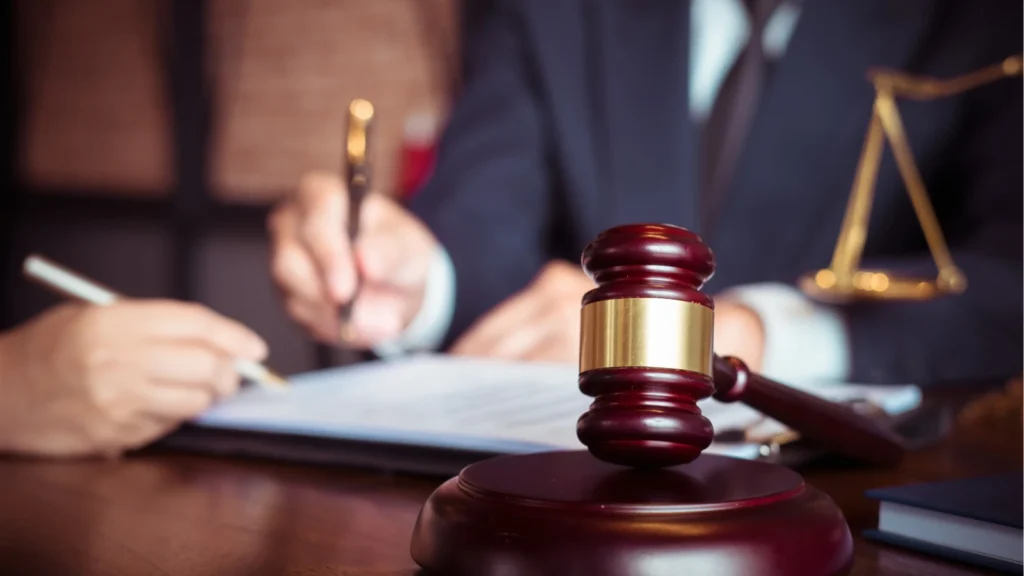 A close-up view of a wooden judge's gavel sitting on a desk while legal professionals sign documents in the background.