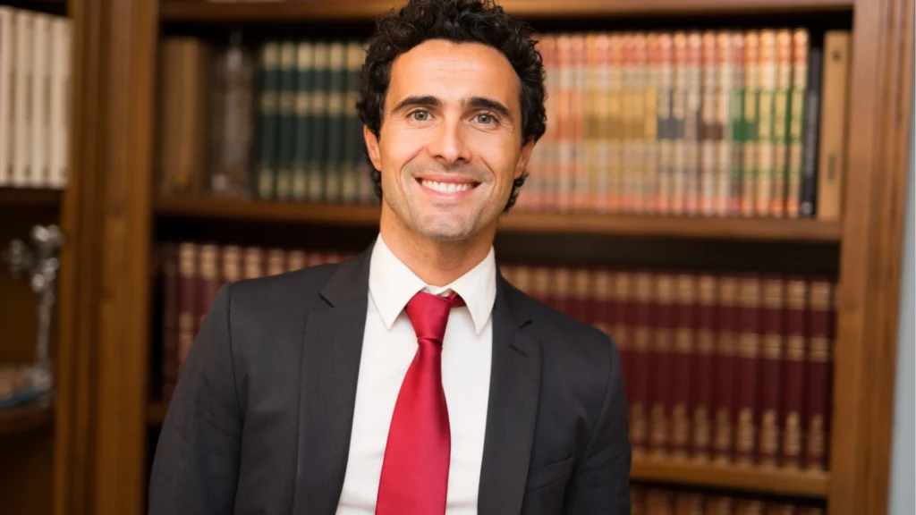 A man wearing a suit and red tie stands in front of bookshelves filled with legal volumes.