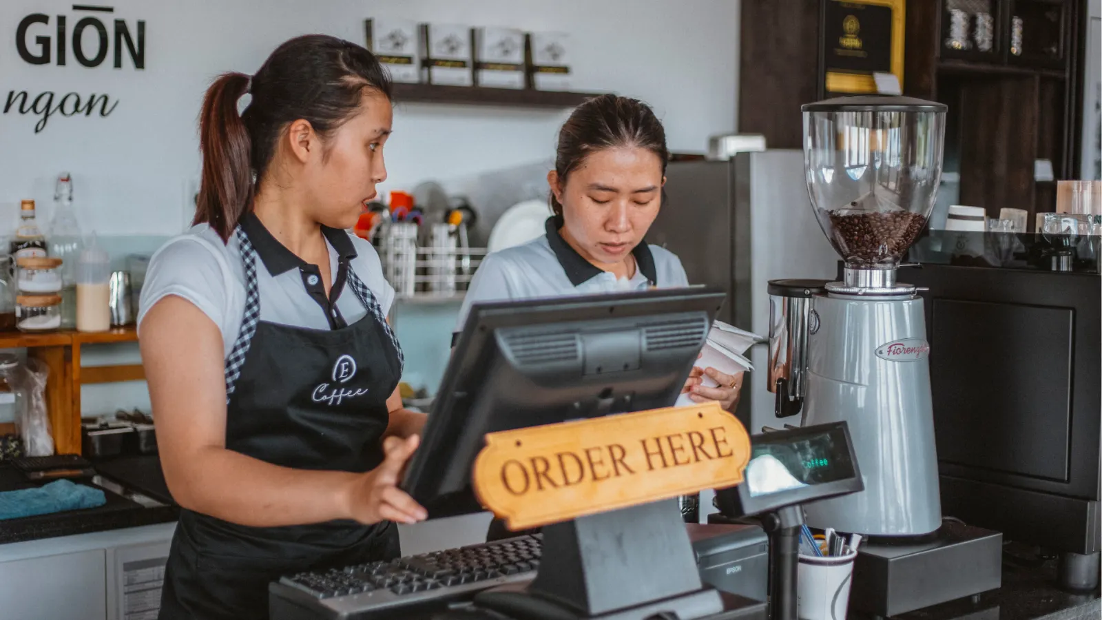 Two coffee shop workers interact at the register counter next to an "Order Here" sign and a coffee grinder.