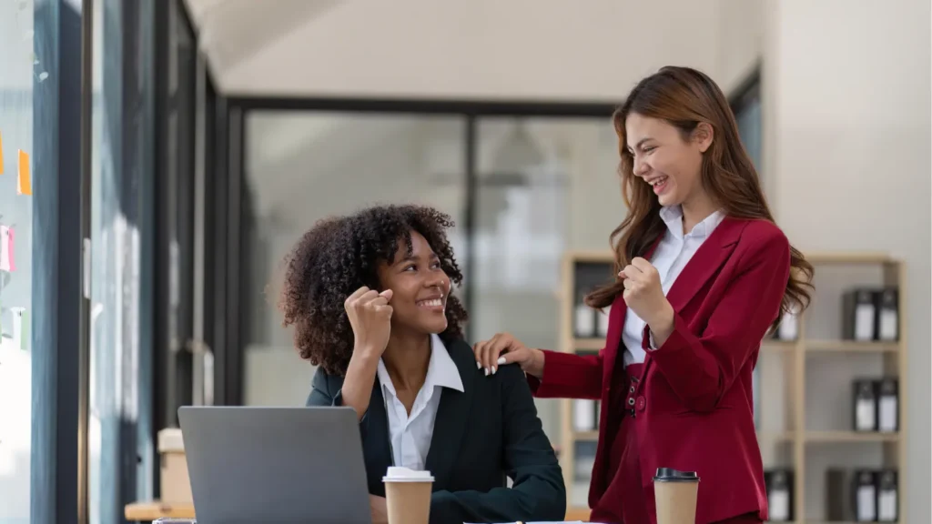 Two professional women in suits smiling and cheering with raised fists at a desk with laptops.