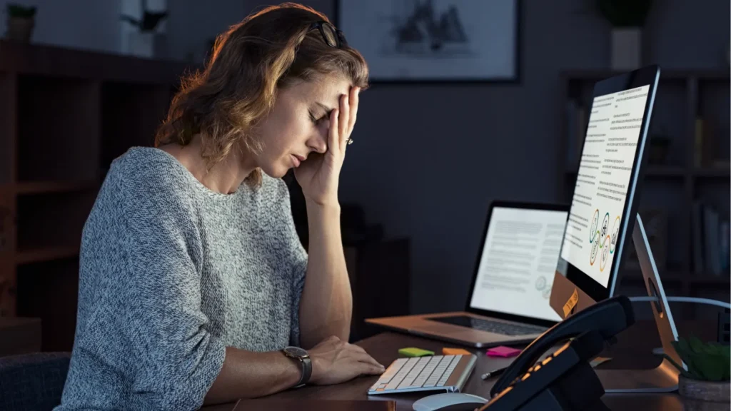 Stressed woman holding her head while working late at a computer in a dark office.