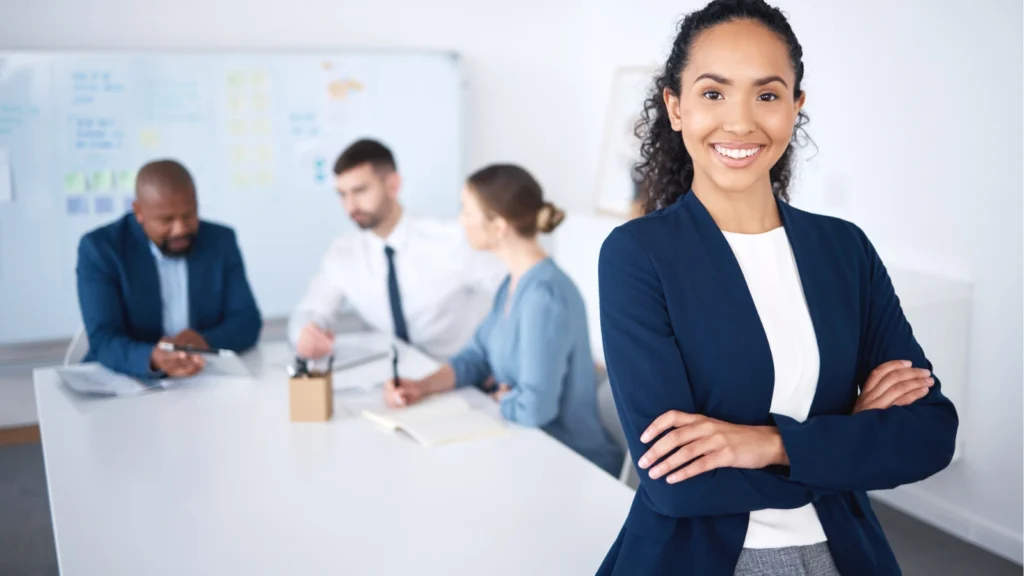 A smiling woman stands in the foreground with crossed arms while her colleagues hold a meeting around a table in the background.