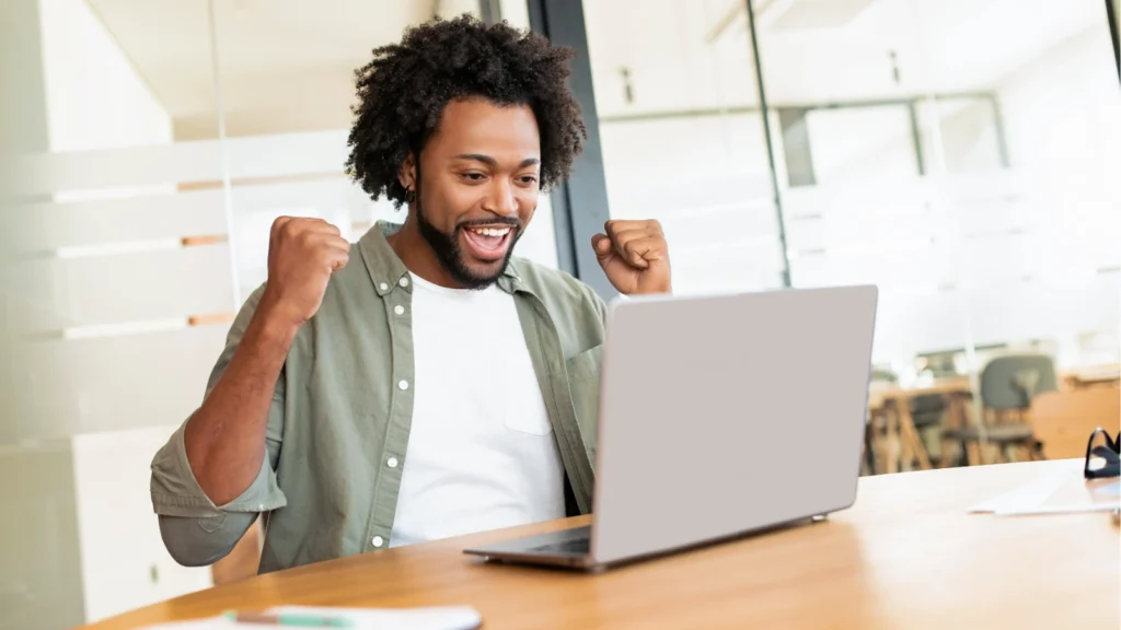 A excited man sits at a wooden desk celebrating with raised fists while looking screen of his laptop.