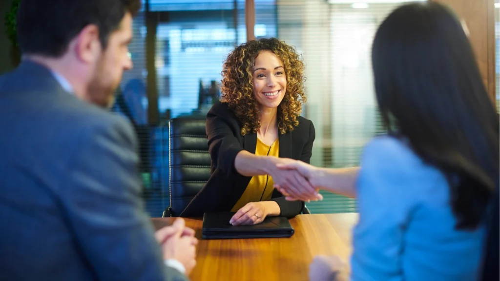 A woman with curly hair smiles as she reaches across a conference desk to shake hands with a client.