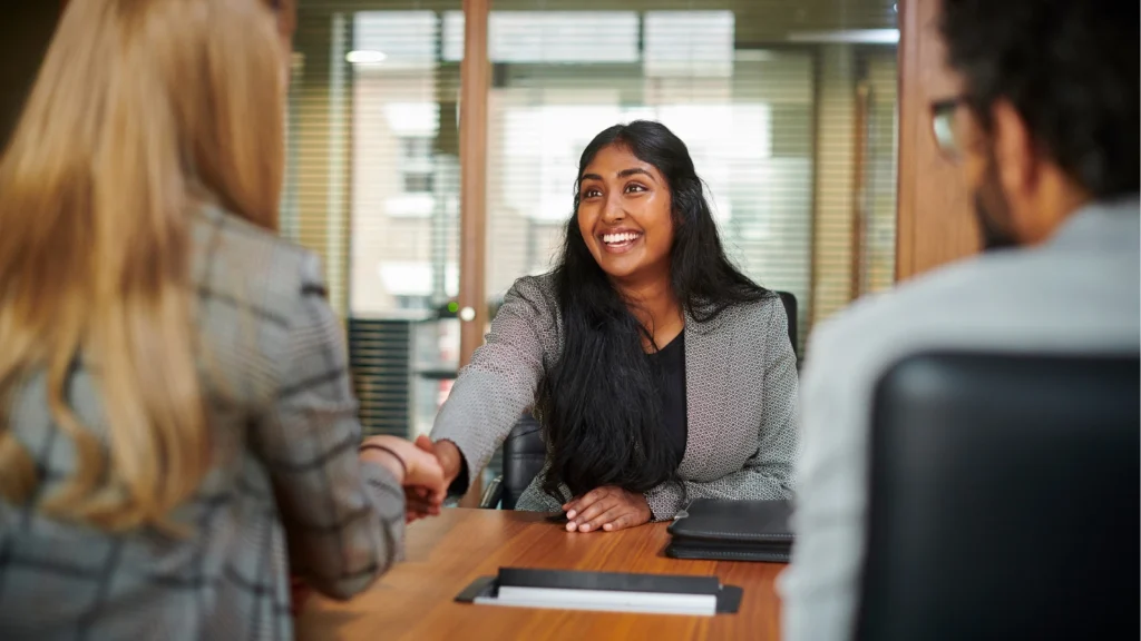 A smiling woman with long dark hair shakes hands across a wooden table during a job interview meeting.