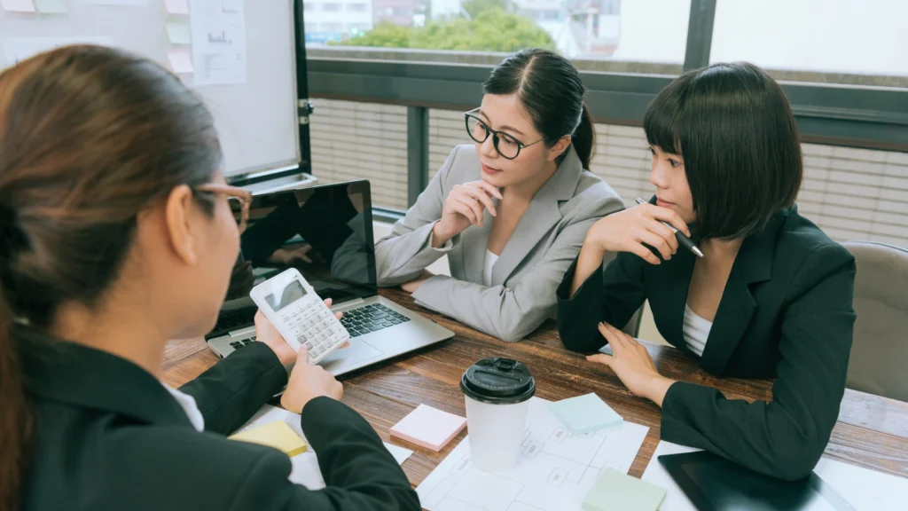 Three business professionals in blazers gather around a table to review financial figures displayed on a calculator and laptop.