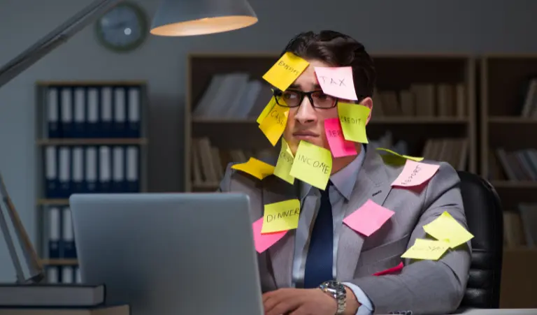 An overwhelmed office worker stares at a laptop, covered in colorful sticky notes, as a desk lamp and shelves frame the busy workspace.