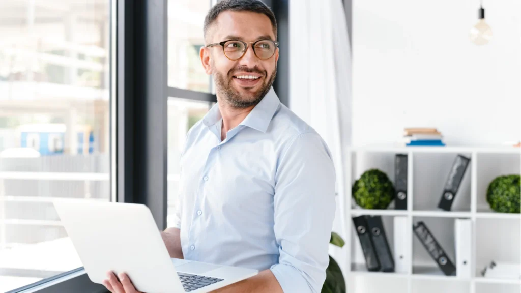 A smiling man in a light blue button-down shirt and glasses holds an open laptop near a window in a bright office.
