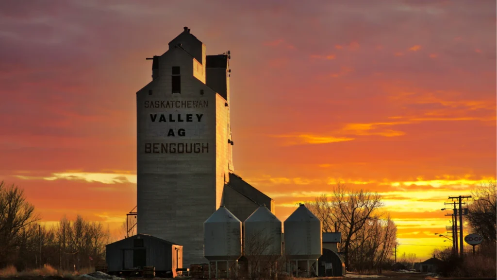 A large wooden grain elevator labeled "SASKATCHEWAN VALLEY AG BENGOUGH" stands against a vibrant orange sunset sky.