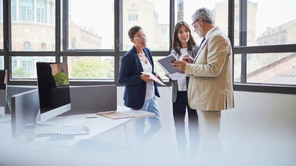 Three business professionals in blazers stand near office windows discussing paper documents together.