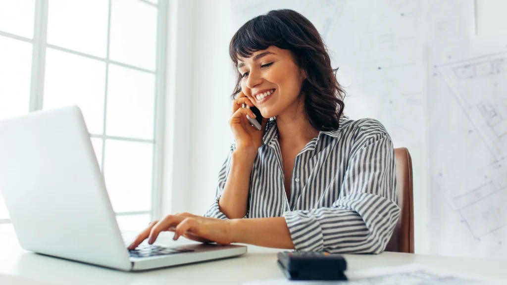 A smiling woman types on a laptop while talking on the phone at a bright office desk.