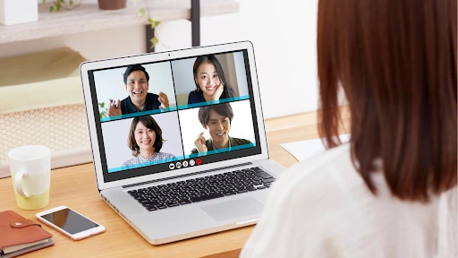 A woman joins a cheerful video conference on her laptop, with four smiling faces on screen; a phone, drink, and notebooks sit on the desk.