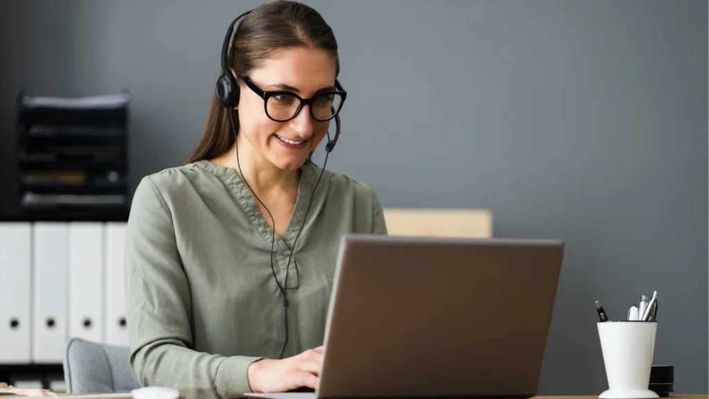 Smiling woman in glasses and a headset works on a laptop at her office desk, with files behind her; she appears focused and approachable.