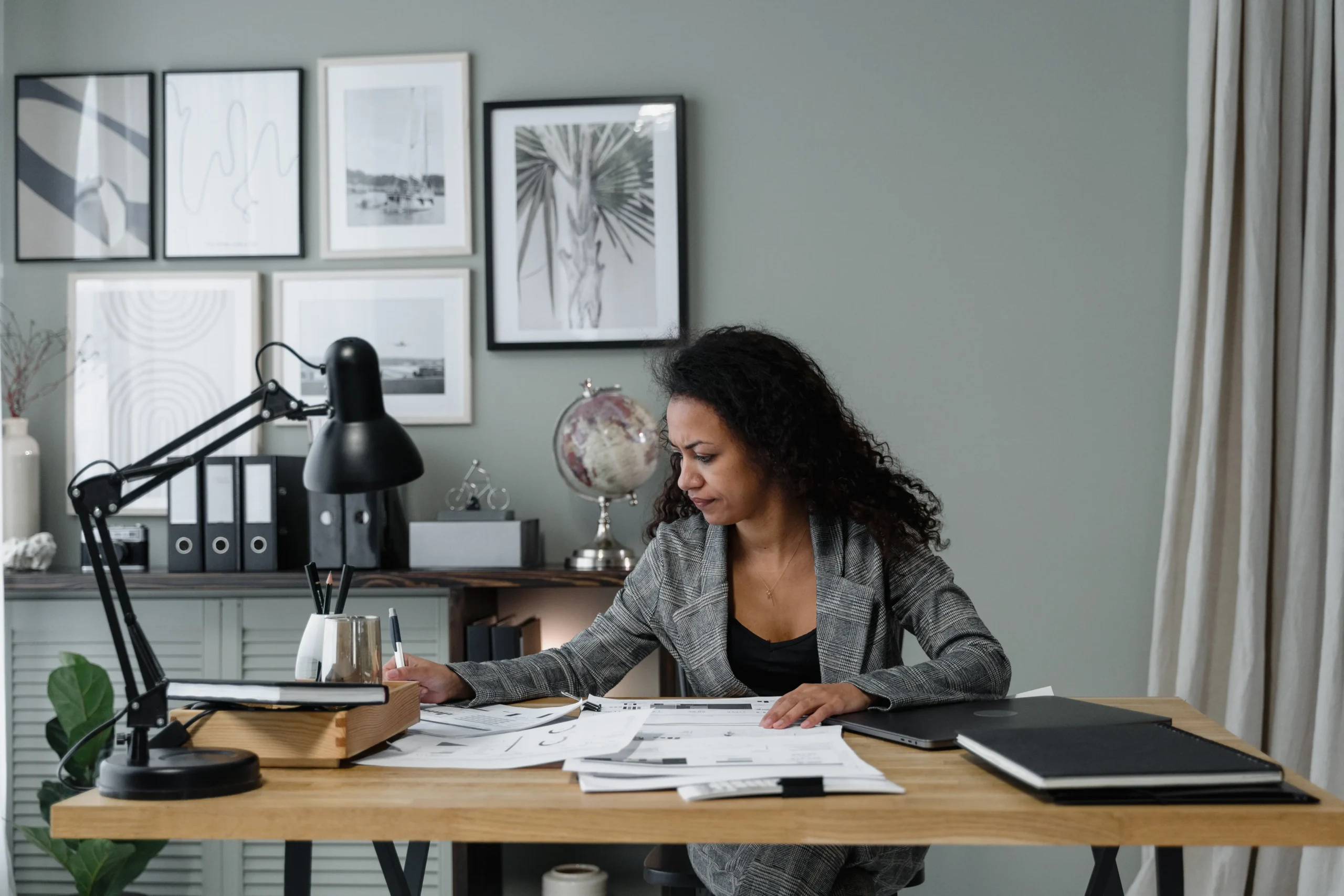 A woman sits at a wooden desk with papers, deep in thought. The room has a calm tone, featuring a globe lamp, framed art, and organized office supplies.