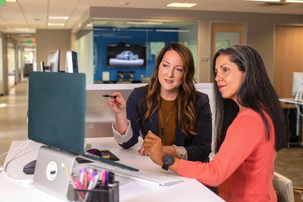 Two women are collaborating at a desk in a modern office, focused on a computer screen. One gestures with a pen, conveying teamwork and discussion.