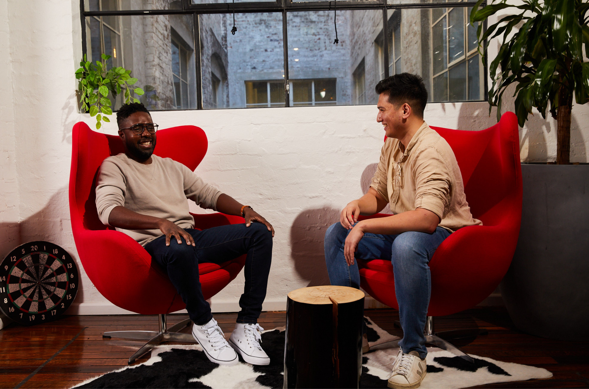 Two colleagues sitting in modern red chairs, smiling and having a relaxed conversation in an industrial-style office space with large windows and plants in the background