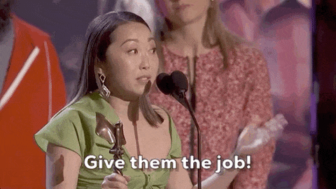 Person speaking into a microphone, holding an award, with text that reads 'Give them the job!
