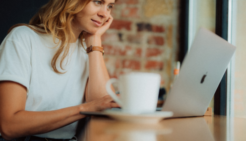 A woman sits in a cafe, looking at her laptop with a mug of coffee next to her