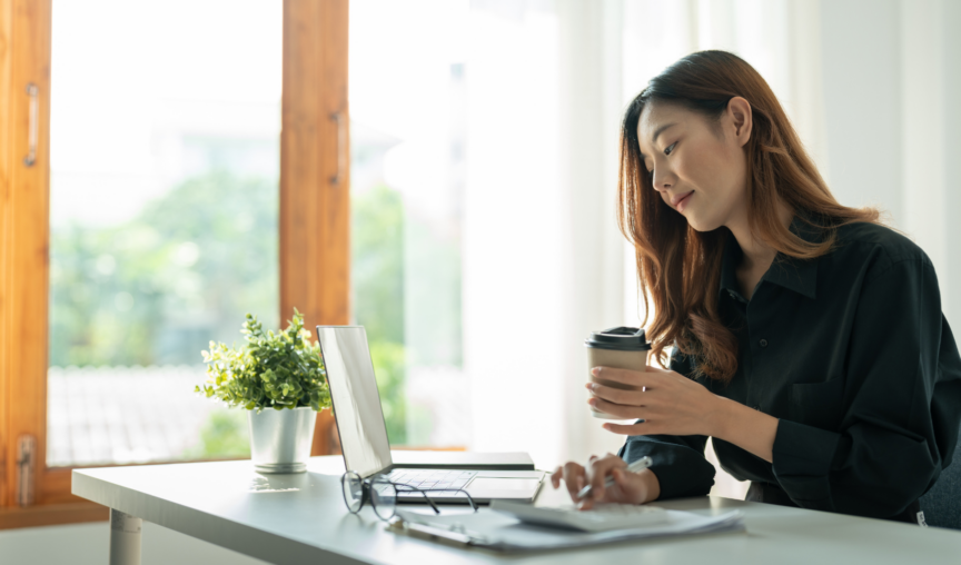 A woman sitting at a desk with a laptop, holding a coffee cup, looking focused while working in a well-lit room