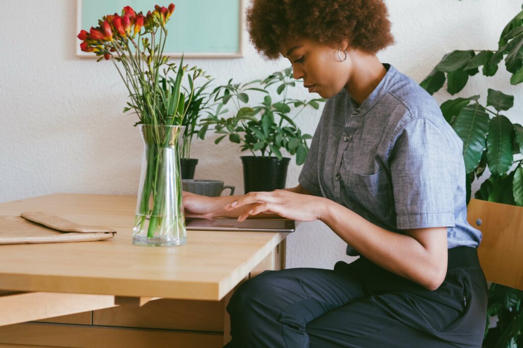 Woman working on a tablet at a wooden table surrounded by indoor plants and a vase of flowers in a cozy home office setting.