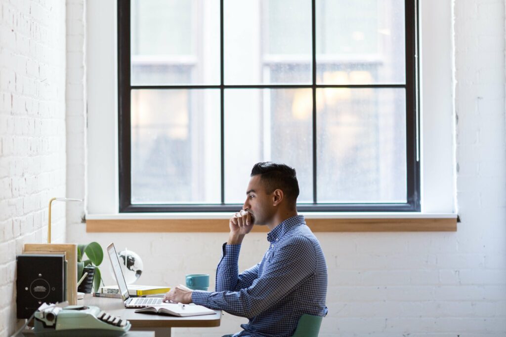 Man sitting at a desk in front of a large window, working thoughtfully on a laptop in a bright, modern office space.