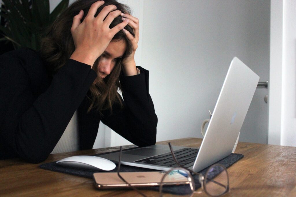 A woman sits at a desk with her hands on her head, appearing stressed while looking at a laptop screen.