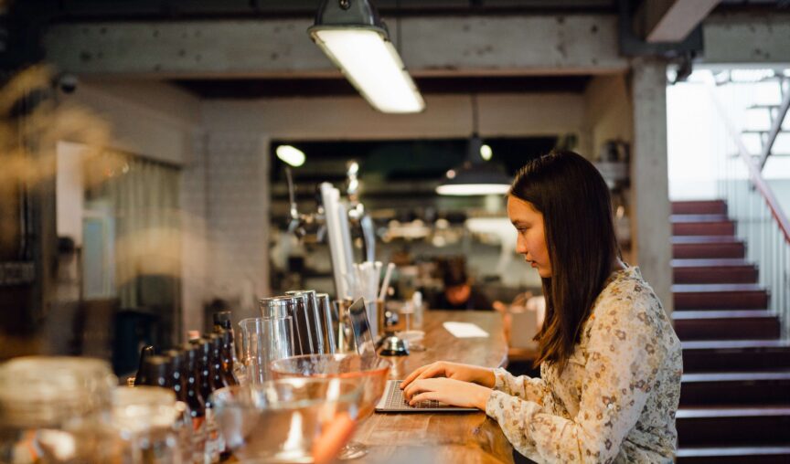 Woman working on a laptop at a bar counter in a cozy, industrial-style cafe or restaurant.