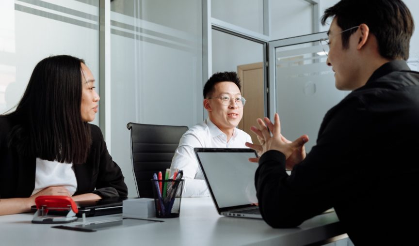 Three colleagues sit around a desk in a modern office, engaged in a discussion with a laptop open in front of them.