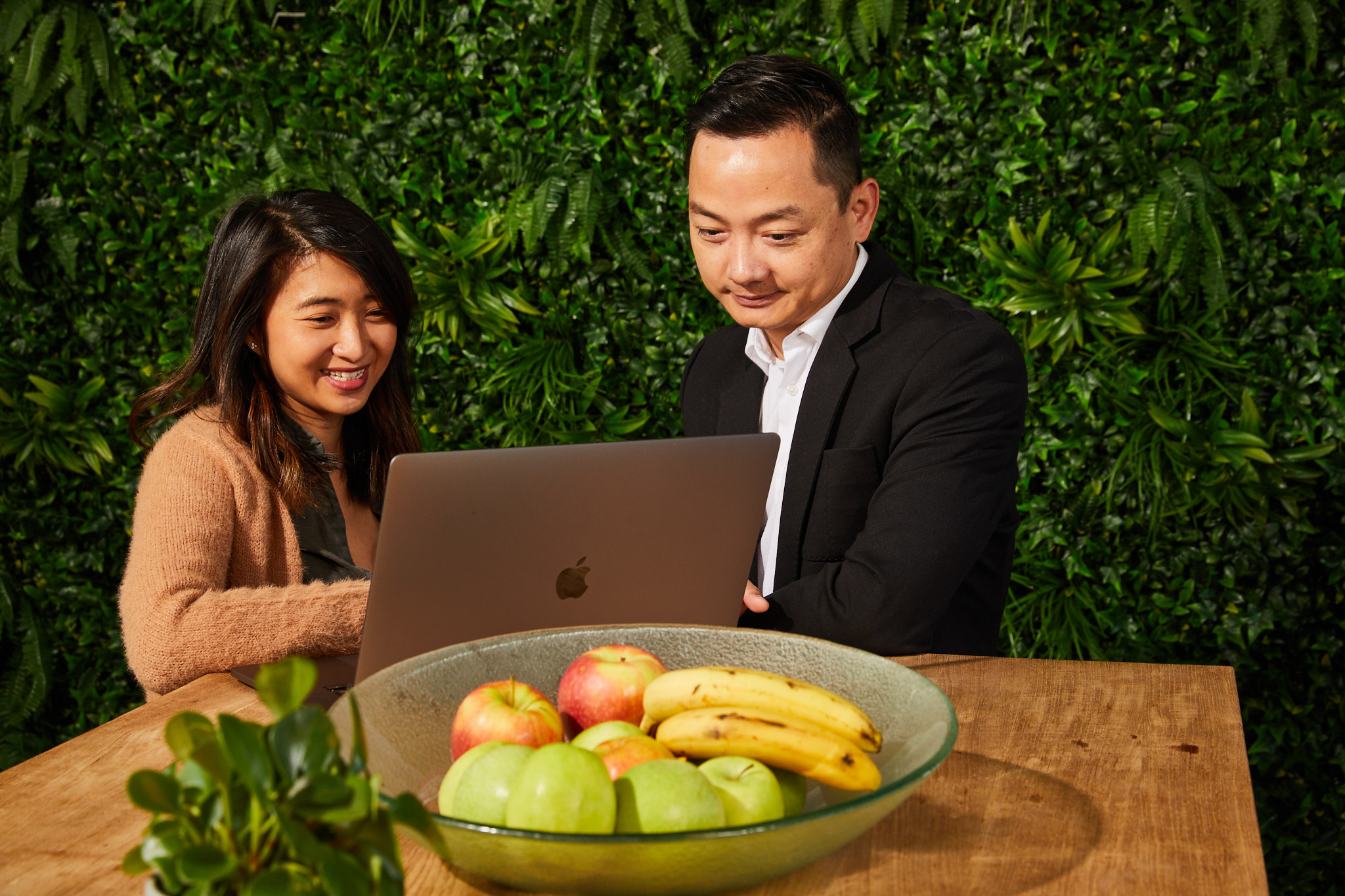 Two people working together on a laptop at a table with a bowl of fruit, set against a green leafy background