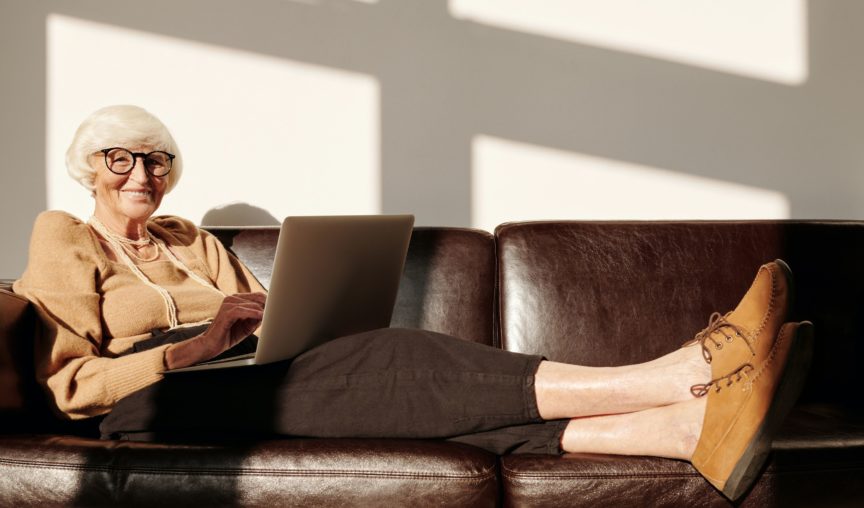 Smiling elderly woman with glasses uses a laptop while relaxing on a leather couch in sunlight.
