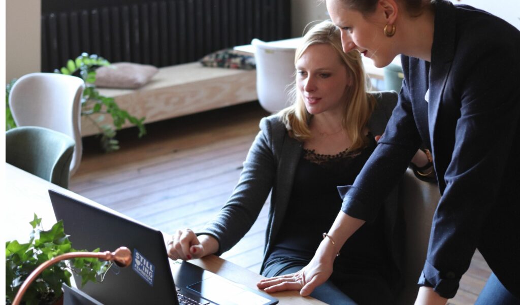 Two women in a modern office setting look at a laptop together, with one seated and the other standing and pointing at the screen.