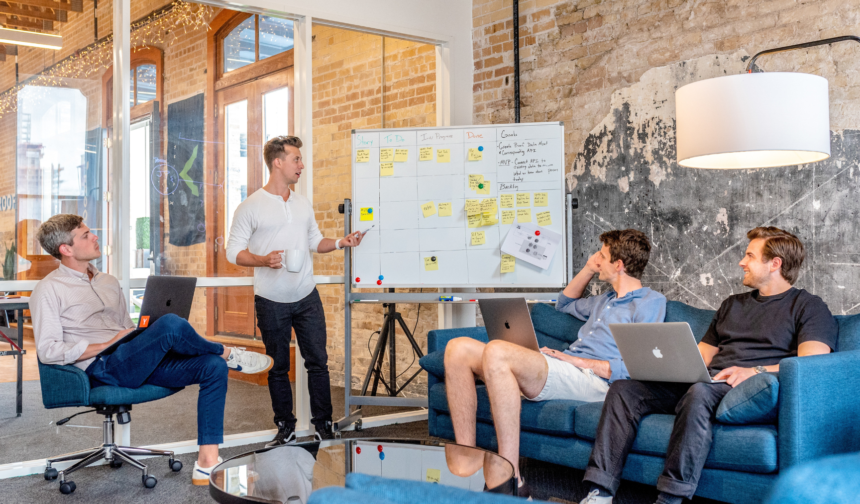 A group of people in a meeting room discuss a whiteboard, while one man leads the discussion