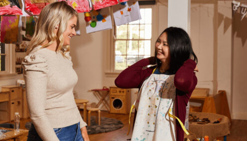 Two women working in a daycare, one is putting on an apron