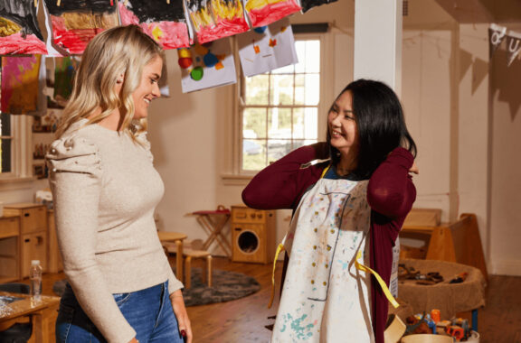 Two women working in a daycare, one is putting on an apron