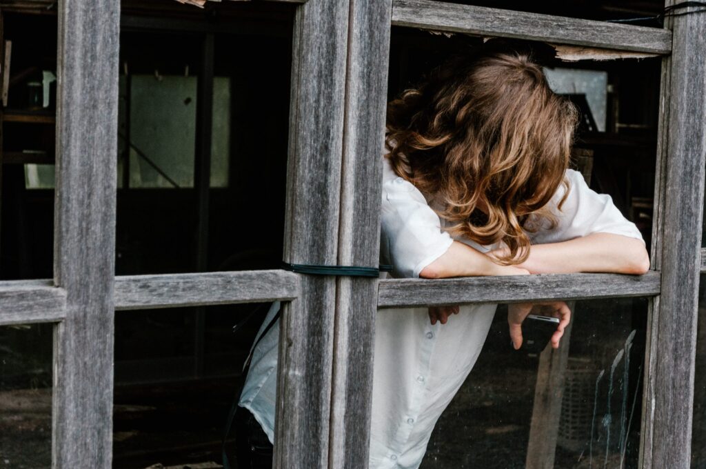 Person leaning out of a rustic wooden window frame with head down and arms resting, conveying a mood of fatigue or contemplation.