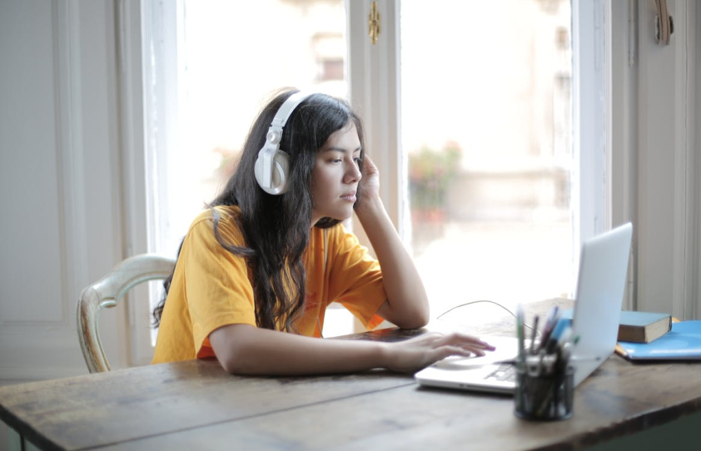 Young woman in headphones looks bored while using a laptop at a wooden desk near a bright window, resting her head on her hand.