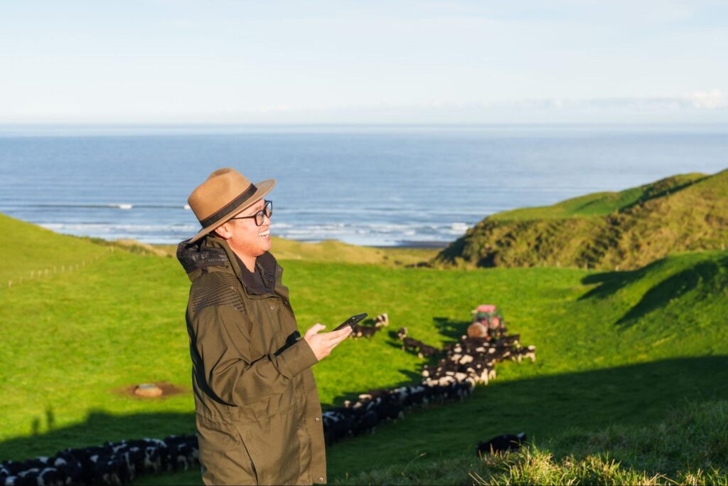Man in hat smiles while holding phone on grassy hillside.

