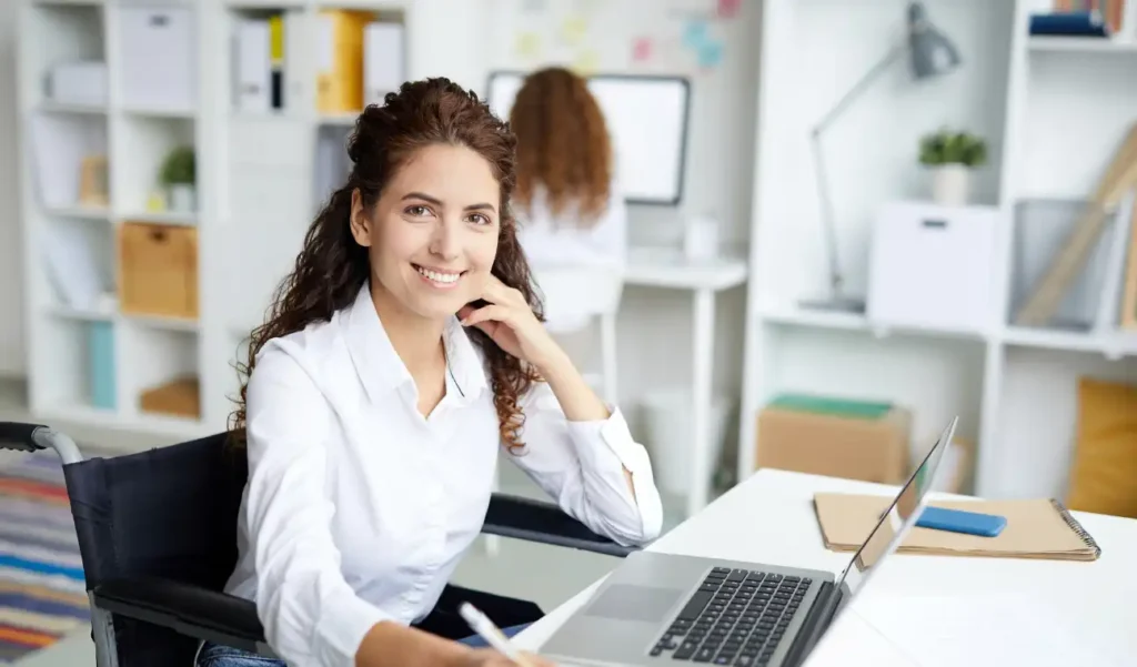 A woman in a white shirt smiles confidently at her desk, using a laptop. She sits in a modern office with shelves and a colleague working in the background.