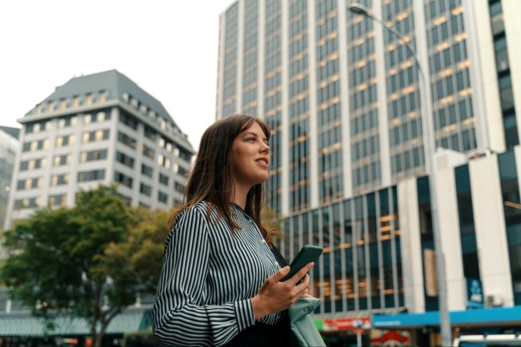 Woman holding phone smiles while standing on city street.