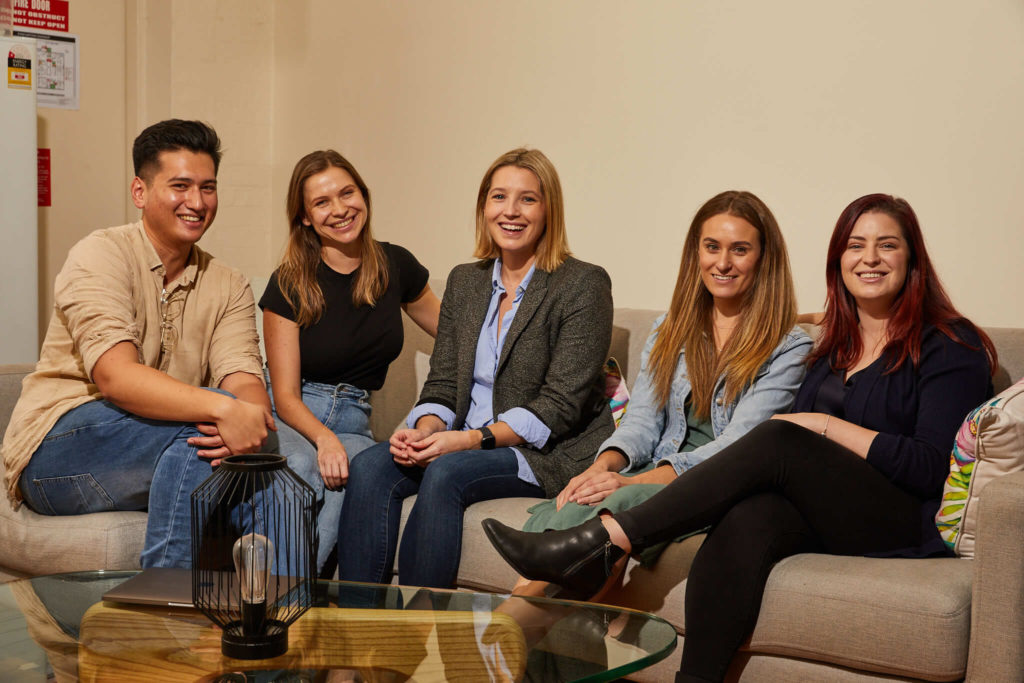 A group of five colleagues sitting on a couch, smiling and relaxed. They are casually dressed, creating a welcoming and friendly atmosphere. A small glass coffee table in front of them holds a decorative lantern and a laptop