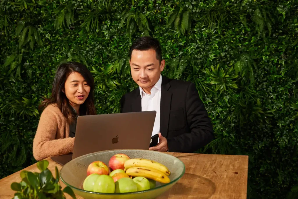 Two people look at a laptop on a wooden table with a bowl of fruit, against a lush green wall. They appear engaged and focused.
