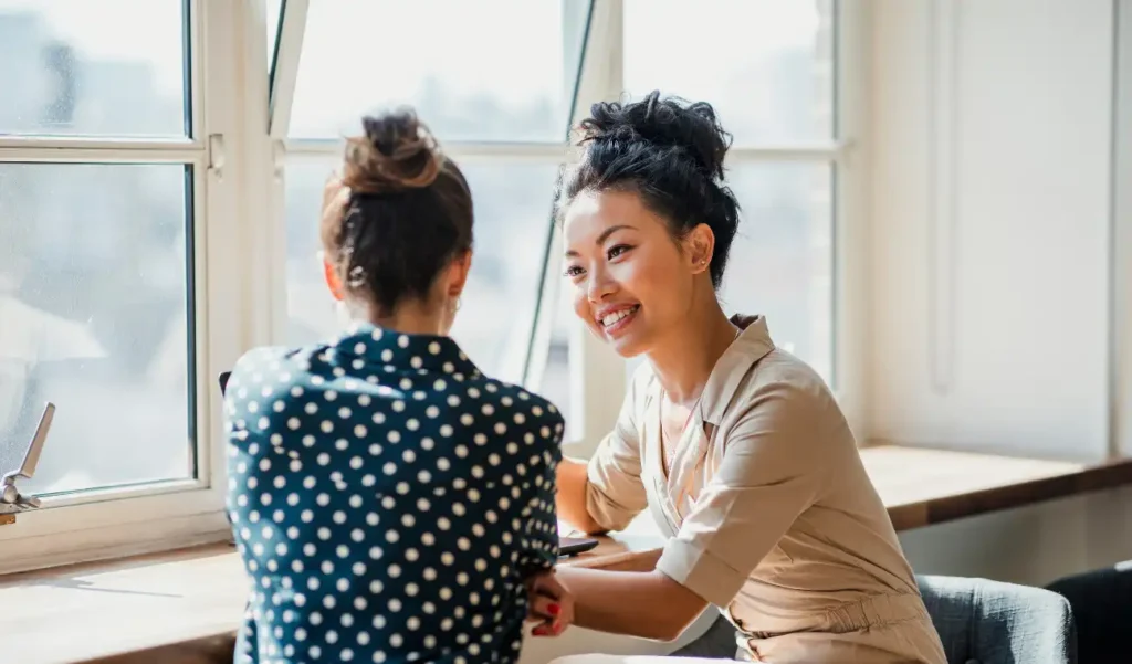 Two women sit by a window in a sunlit room, engaged in an animated conversation. One wears polka dots, the other beige, both smiling warmly.
